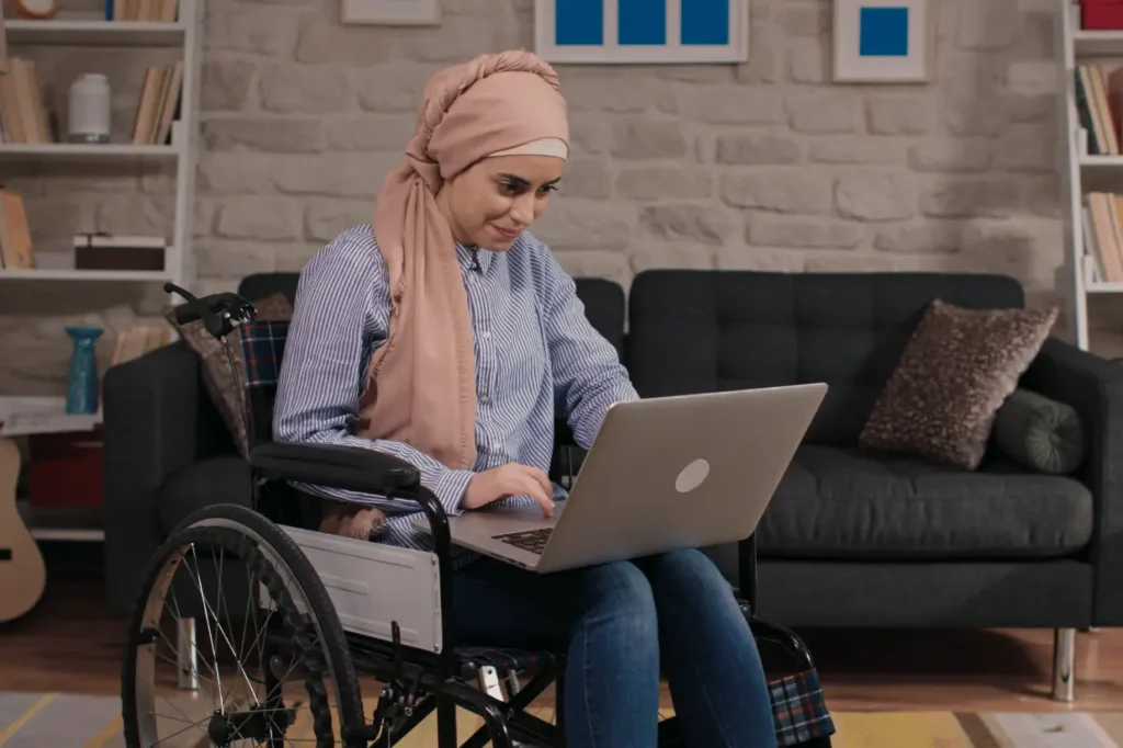 Muslim woman in wheelchair sitting in lounge room typing on laptop
