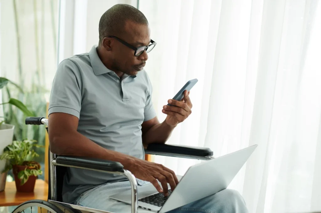 Disabled man in wheelchair typing on laptop while talking on mobile phone